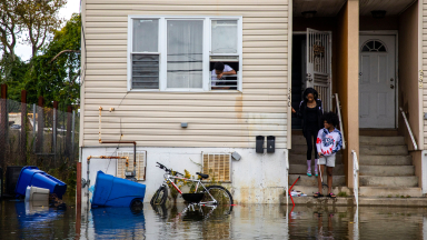 A flooded street in front of the home of Danielle Smith who is standing with her preteen daughter on a set of exterior concrete steps that are nearly submerged. A man in a white T-shirt looks out of a first floor window.